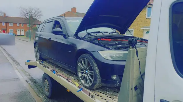 A car on a recovery truck bed with its bonnet up and jump leads on the battery